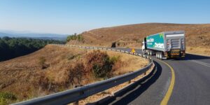 Ciężarówka jedzie po drodze, wokół widać wypłowiałe łąki, a nad nią jest błękitne bezchmurne niebo.